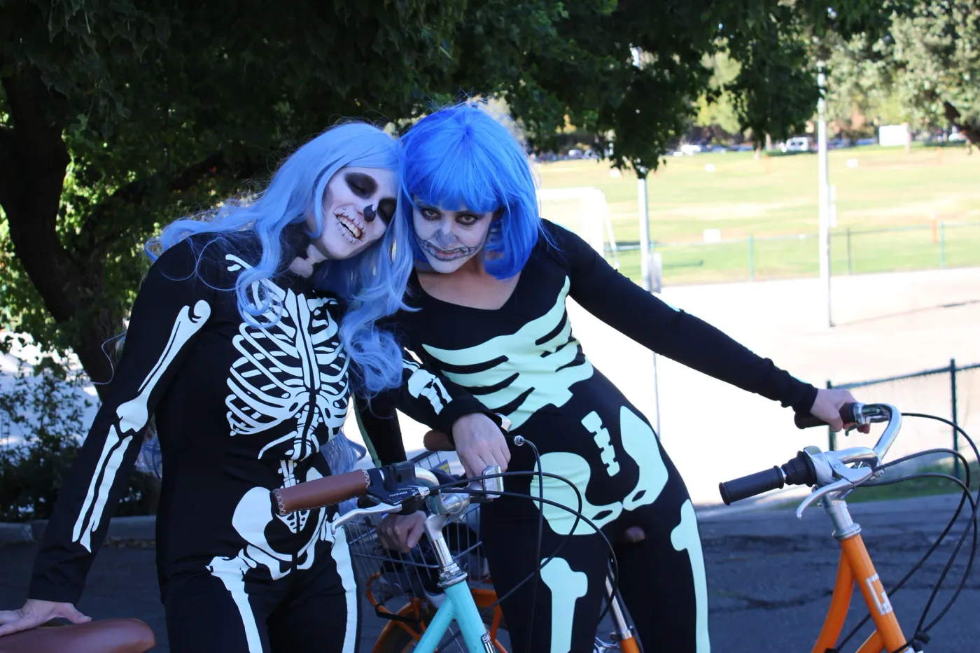 Two riders in blue wigs and skeleton face paint leaning together beside their bicycles.